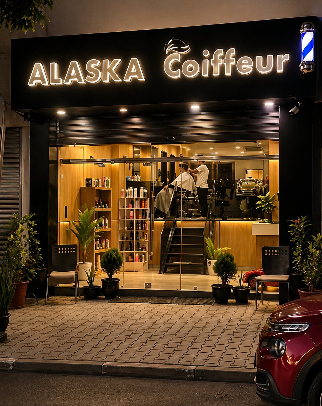 Storefront of Alaska Coiffeur hair salon at night with illuminated signage, warm-lit interior displaying styling stations and products, potted plants flanking entrance, parked car visible.