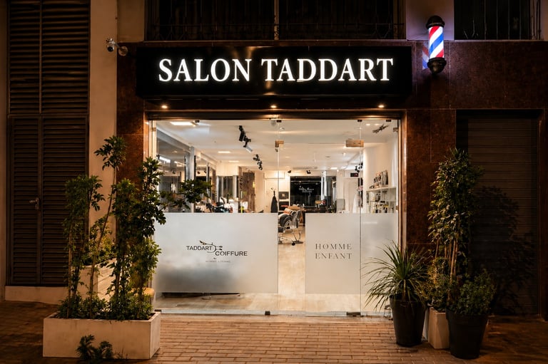 Nighttime storefront of Salon Taddart with illuminated signage, glass entrance doors, and potted plants flanking the entrance